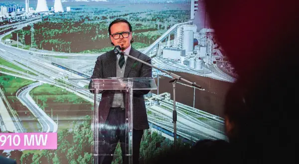 Mężczyzna przemawia na tle wizualizacji elektrowni i autostrady. Na ekranie widnieje napis "910 MW", symbolizujący zwiększenie mocy przyłączeniowej Tauron.