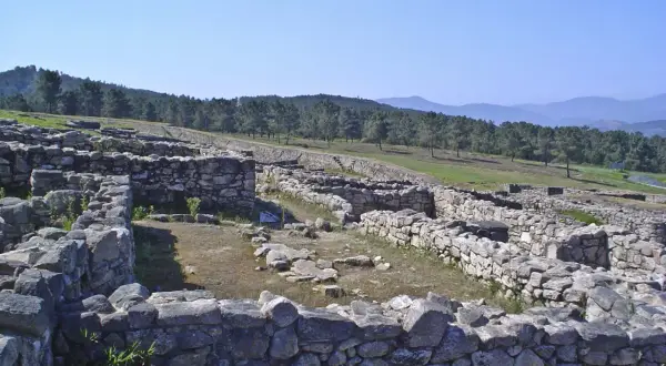 Ruinas del castro de San Cibrao de Las, con muros de piedra y un paisaje boscoso al fondo bajo un cielo azul.