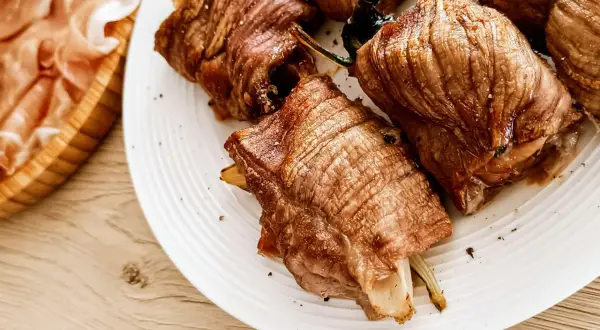 Involtini di carne succulenti, cotti alla perfezione, serviti su un piatto bianco. A lato, fette di prosciutto crudo in un cestino di legno.