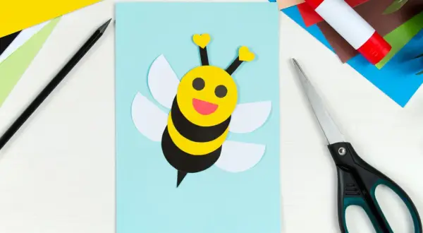 Ein süßes Biene basteln-Projekt auf hellblauem Papier. Bunte Bastelmaterialien, Schere und Klebestift liegen bereit.