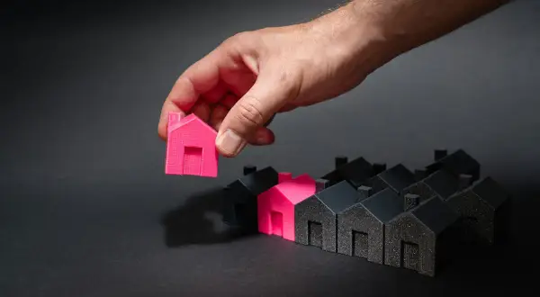 Mano che sceglie una casetta rosa tra tante nere. Un'idea di permuta immobiliare con costruttore.