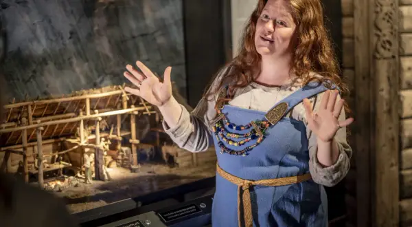 En kvinna i vikingakläder berättar vid en modell av en vikingagård på ett museum i Stockholm.
