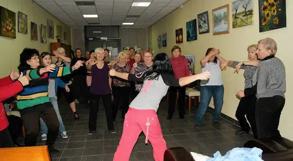 Grupa seniorów ćwiczy w sali Domu Seniora Zielony Zakątek. Wszyscy uśmiechnięci, w ruchu, w tle obrazy.