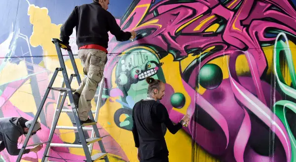 Drei Künstler sprühen bunte Graffiti auf eine Wand, einer auf einer Leiter.