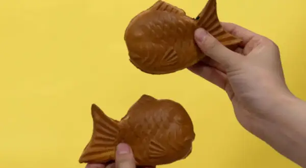 Zwei goldbraune Taiyaki, beliebte japanische Desserts in Fischform, werden von Händen auf gelbem Hintergrund gehalten.