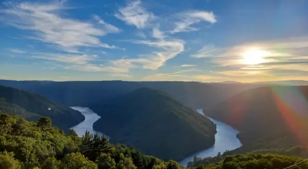 Un río serpentea entre montañas verdes, bajo un cielo azul con nubes finas. El sol se pone, tiñendo el horizonte de dorado. Parece el cabo do mundo.