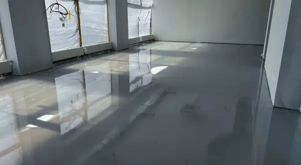 Lśniąca podłoga z żywicy w pomieszczeniu w budowie. Widać odbicia okien i instalacji.