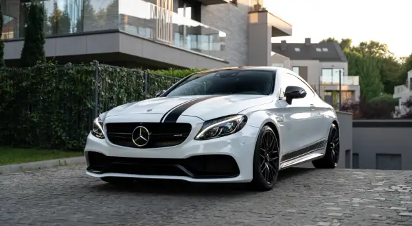 Biały Mercedes-AMG C63 Coupe z czarnymi pasami na masce, zaparkowany na brukowanej drodze przed nowoczesnym budynkiem.