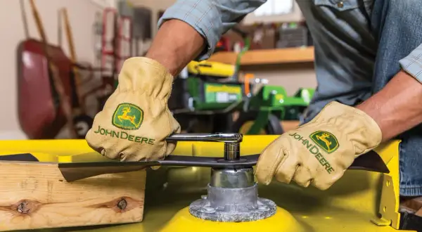 Gloves on, a person uses a socket wrench to remove a mower blade, preparing to sharpen it.