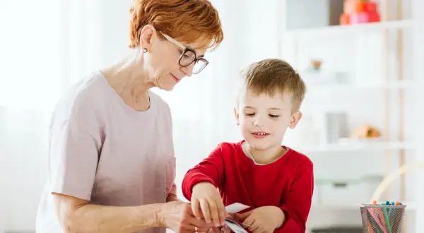 Babcia i wnuczek tworzą coś razem. Dziecko z zaciekawieniem przygląda się swoim ruchom, a jego manieryzmy ruchowe są widoczne w skupieniu.