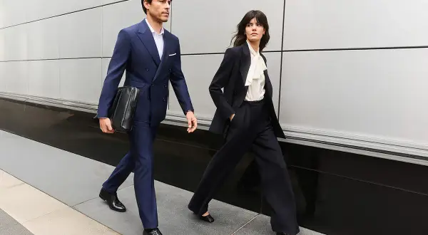 Para idąca ulicą w eleganckich strojach, idealnych na dress code business casual. Mężczyzna w granatowym garniturze, kobieta w ciemnym kostiumie.
