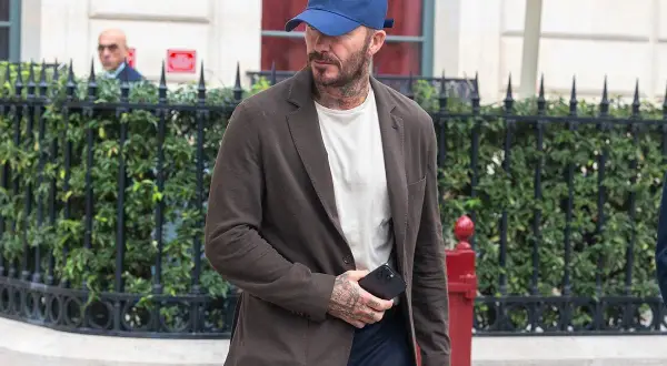 Uomo con abbinamento blu e marrone: blazer marrone, t-shirt bianca, cappellino blu e pantaloni scuri.