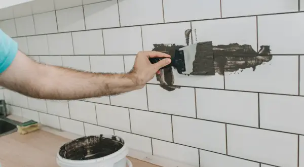 Ręka nakłada ciemną fugę na białe płytki, pokazując, jak zmienić kolor fugi na ciemniejszy. Wiadro z fugą stoi obok.