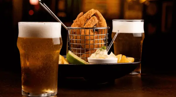 Due bicchieri di birra bionda ghiacciata accompagnano un cestino di anelli di cipolla fritti, patatine e una salsa cremosa.