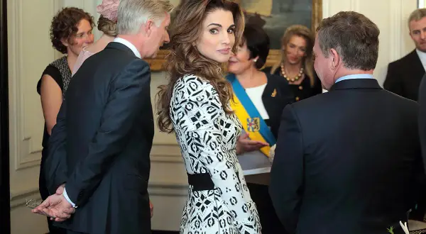 Regina Rania di Giordania, elegante in abito bianco e nero, partecipa a un evento. Sullo sfondo, un quadro e altre personalità.