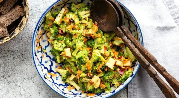 Frischer Brokkoli Salat mit Apfelstücken und Karottenraspeln, serviert in einer blauen Schüssel mit Holzkochlöffeln.
