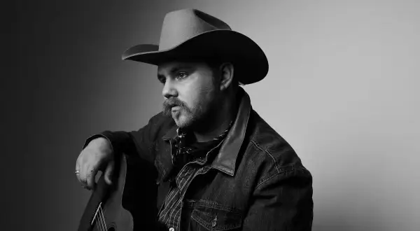 Ernest Keith Smith, a thoughtful country musician, poses with his guitar, wearing a cowboy hat and denim jacket.