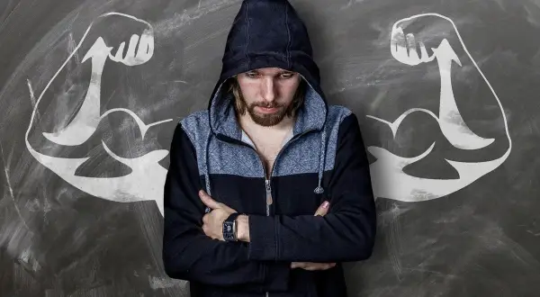 Chłopak w bluzie z kapturem, z ramionami skrzyżowanymi na piersi, stoi przed tablicą z rysunkami bicepsów. Idealny materiał na komplementy dla faceta, który czuje się silny.