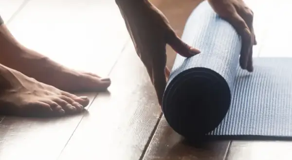 Pieds nus sur un parquet, une main déroule un tapis bleu pour une séance de sport à la maison.