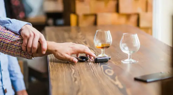 Ręka powstrzymuje drugą przed sięgnięciem po kluczyki do samochodu. Na stole stoją kieliszki z alkoholem, sugerując ryzyko utraty prawa jazdy za alkohol.