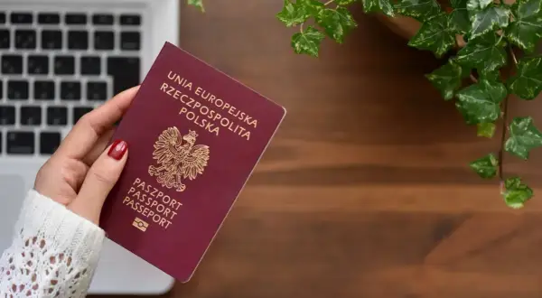 Dłoń trzyma polski paszport na tle laptopa i roślin.