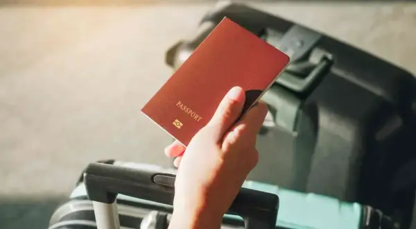 Dłoń trzyma paszport, obok walizka. Problem z wyrabianiem nowego dokumentu z powodu braku starego paszportu.