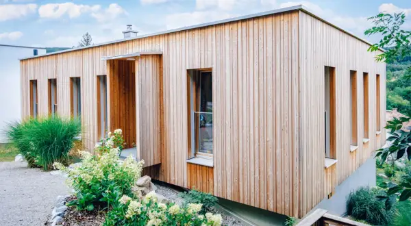 Modernes Haus mit einer Mischfassade aus Holz und Putz. Vertikale Holzverkleidung umrahmt schmale Fenster. Grüne Pflanzen schmücken den Eingangsbereich.