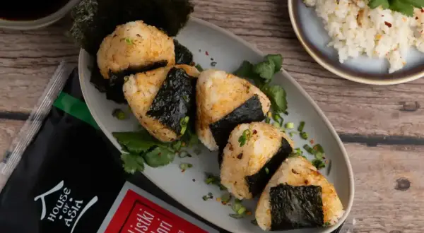 Pięć onigiri z ryżem, udekorowanych listkami nori i szczypiorkiem, obok opakowania nori i miseczki z ryżem.
