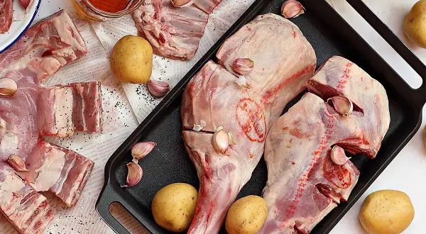 Pierna de cordero cruda con patatas, ajos y aceite de oliva, lista para asar.