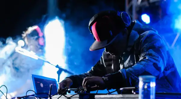 Un techno dj con cuffie e cappellino rosso mixa musica su un palco illuminato da luci blu e fumo.