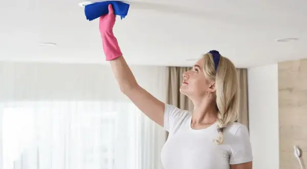 Une femme blonde en gant rose nettoie un luminaire au plafond avec un chiffon bleu. Elle montre comment nettoyer un plafond.