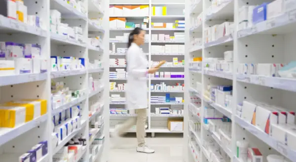 Farmaceutka w białej kitce porusza się między regałami pełnymi leków, szukając potrzebnego produktu. W aptece, gdzie panuje porządek, można znaleźć wiele preparatów, w tym te z grupy polpril.