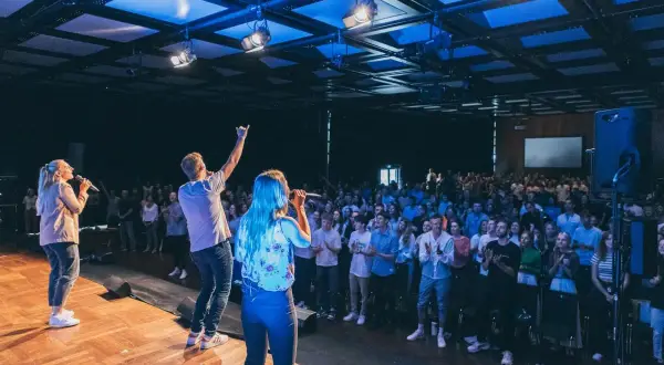 Junge Leute singen auf einer Bühne vor einer großen Menge. Ein Mann hebt die Hand, als ob er eine Botschaft verkündet. Eine moderne Kirche, die urbanes Leben und Gemeinschaft vereint.