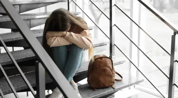 Smutna dziewczyna siedzi na schodach, przytulając kolana. Obok niej leży plecak. Martwi się o warunkowe przejście do następnej klasy.