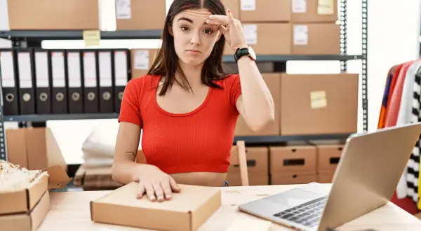 Mujer frustrada revisando el inventario online, rodeada de cajas y carpetas.