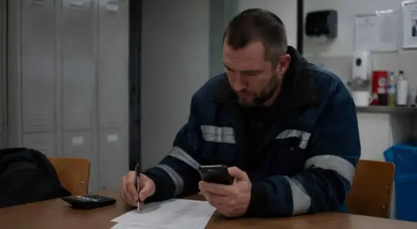 Pracownik w roboczym ubraniu sprawdza telefon, pisząc coś na papierze. Może to być rozliczenie za dodatek za drugą zmianę.