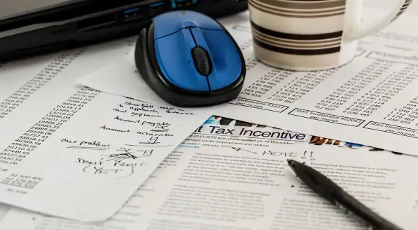 Biurko z laptopem, myszką, kubkiem i dokumentami. Na papierach widać tabelę, notatki "our problems x-y", "DON'T PANIC (YET)" oraz nagłówek "Tax Incentive".