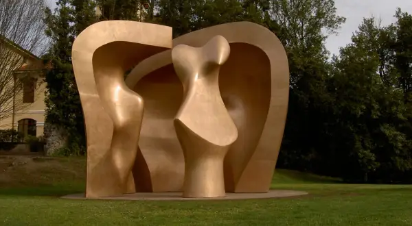 Escultura abstracta dorada en el parque de los pueblos de Europa, con formas curvas y orgánicas, rodeada de césped y árboles frondosos.