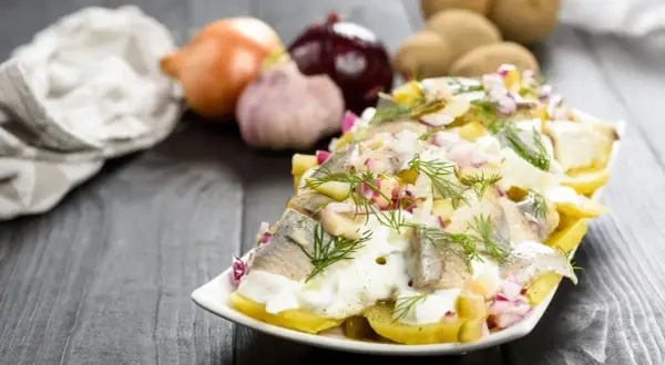 Pyszna sałatka ziemniaczana ze śledziem, udekorowana koperkiem i czerwoną cebulką. Obok leżą ziemniaki, cebula i czosnek.