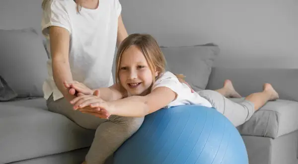 Uśmiechnięta dziewczynka ćwiczy na piłce rehabilitacyjnej, poprawiając swój **tonus mięśniowy** pod okiem mamy.