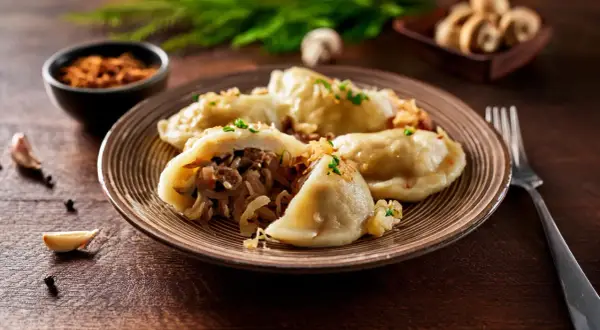 Talerz pełen pysznych, domowych pierogów z kapustą i grzybami, posypanych szczypiorkiem. Obok czosnek i pieprz.