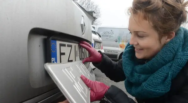 Kobieta w czerwonych rękawiczkach montuje tablicę rejestracyjną. To ważny etap rejestracji samochodu.