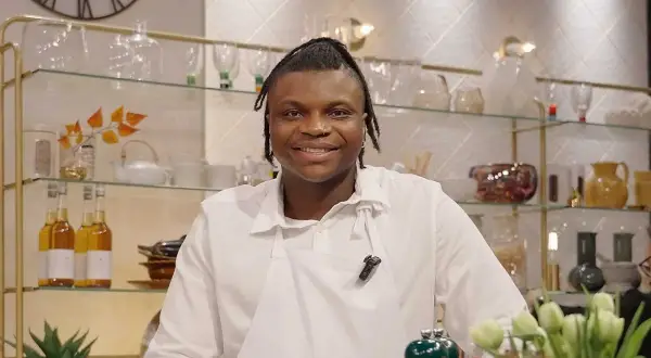 Kelvin Wokomaty, en glad kock i vitt förkläde, ler mot kameran. Bakom honom syns hyllor med flaskor och dekorationer.