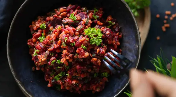 Ein herzhafter Linsen Rote Bete Salat, garniert mit Petersilie, wird mit einer Gabel aus einer dunklen Schüssel genommen.