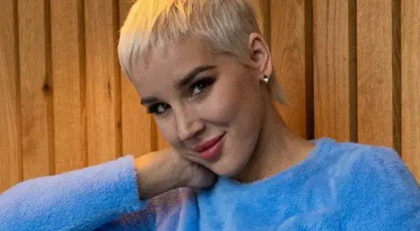 Kobieta z krótkimi blond włosami w niebieskim swetrze opiera głowę na dłoni.
