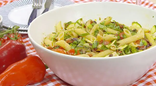 En stor skål med vegansk pastasallad med penne, zucchini, tomater och oliver. Serveras på en rödvitrutig duk.