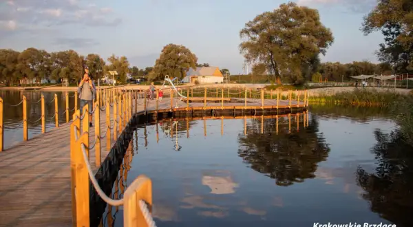 Drewniany pomost nad wodą w największym parku w Krakowie. Ojciec z dzieckiem na ramionach spaceruje po pomoście.