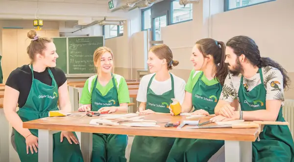 Fünf junge Menschen in grünen Schürzen lachen und arbeiten an einem Holztisch. Hier beginnt ihre kostenfreie Ausbildung Ergotherapie.