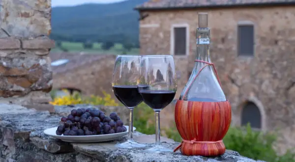 Dwa kieliszki czerwonego wina, kiść winogron i butelka w koszyczku, na tle kamiennego muru i krajobrazu regionu Chianti.