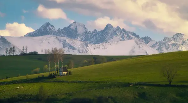 Zielone wzgórza, mały biały domek z drzewami i ośnieżone góry w tle.